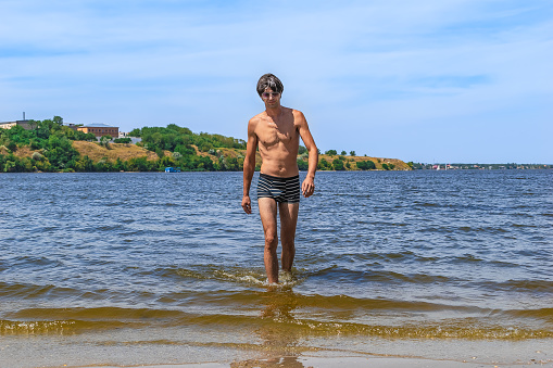 A young slender man in swimming trunks and sunglasses walks out of the river on Strilka beach in Mykolaiv, Ukraine. Rest of people at the confluence of the rivers Inhul and Pivdennyi Buh