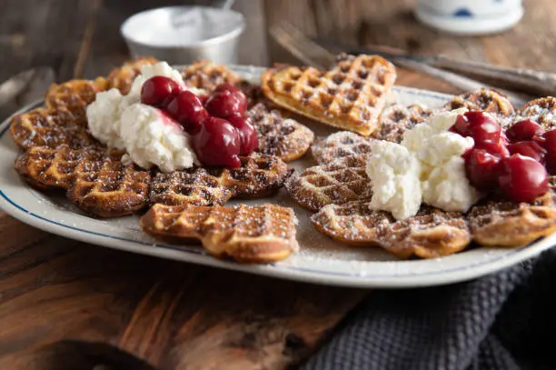 Traditional german dessert with fresh and homemade heart shaped waffles. Topped with whipped cream and sour cherries. Served on rustic and wooden table background. Front view and closeup