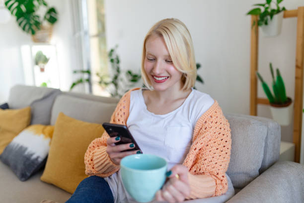 istock Attractive smiling woman using smart phone while sitting on the sofa at home. Communication and coziness concept. 1387033893