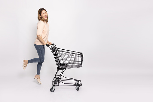 Full length portrait of young Asian woman jumping and pushing an empty shopping cart or shopping trolley isolated on white background