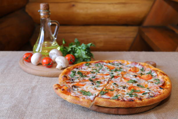 Traditional pizza with salami and champignons in front of a cutting board with mushrooms, tomatoes, jug of oil and bunch of greens against a log wall.