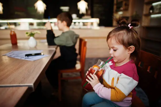 Photo of Funny baby girl sitting in cafe and drink juice lemonade.