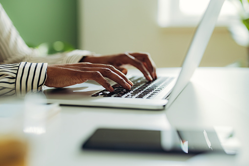 Unrecognizable African American woman typing business report on a laptop keyboard while sitting at office desk.