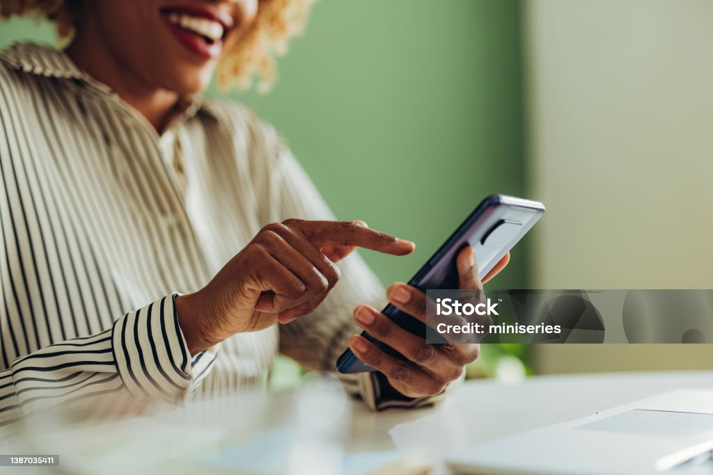 Close Up Photo of Woman Hands Using Mobile Phone in the Office An anonymous African American woman typing text message on her smartphone while sitting at office desk with laptop computer. People Stock Photo