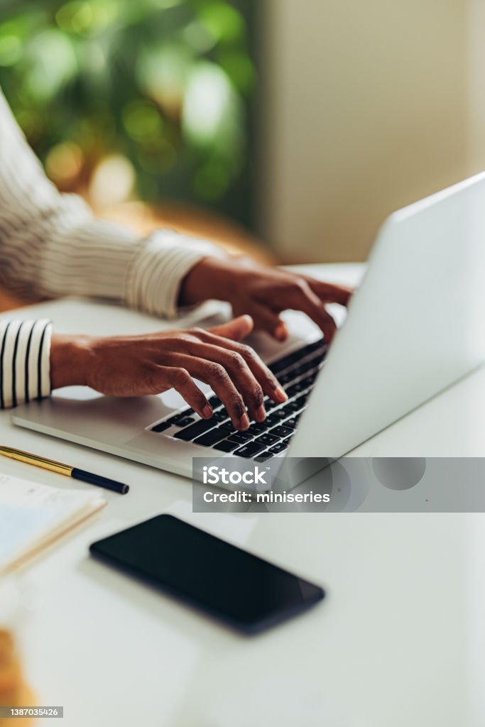 Close Up Photo of Woman Hands Using Laptop Computer in the Office An anonymous African American woman typing business report on a laptop keyboard while sitting at office desk with mobile phone, notebook and pen. Hand Stock Photo