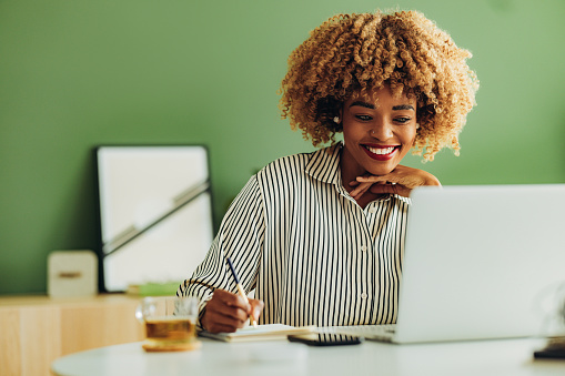 Smiling African American woman reading business report on a laptop and writing notes in notebook while sitting at office desk.