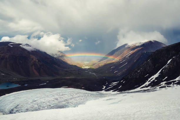 istock Rainbow over a mountain valley. Gloomy scenery with bright rainbow above glacier in mountain valley. Top view to colorful rainbow and low clouds in mountains in rainy and sunny weather. 1387042766