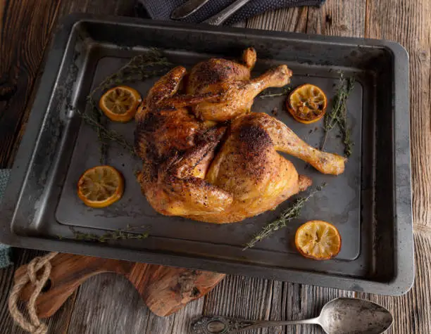 Delicious flat roast whole chicken. Cooked with lemon and thyme. Served in a roasting pan on dark wooden table background. Closeup and overhead view