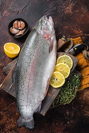 Whole trout, raw fish on a wooden board with thyme and lemon. Dark background. Top view.