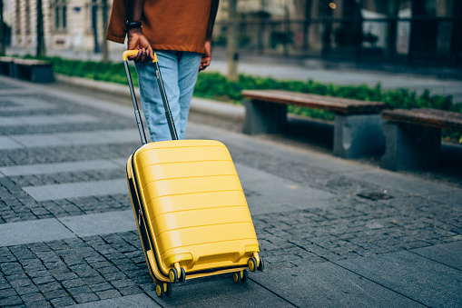 Cropped shot of young afro-american man with suitcase walking on the street in foreign city. Photo of a young man walking down the street and pulling a yellow suitcase. Man traveler walking alone with suitcase bag. Handsome businessman on a business trip. Travel Concept.