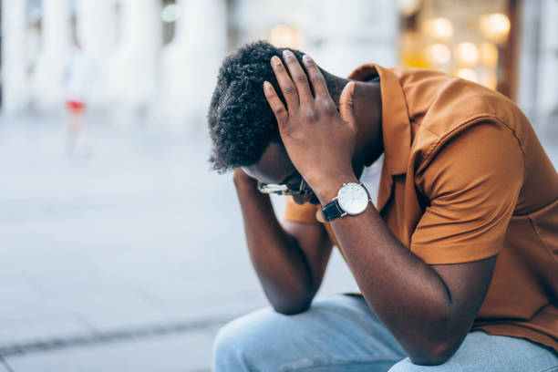 Depressed young man outdoor in the city. Shot of a stressed young afro-american man sitting on bench at the street and holding head with hands. Portrait of black man having a headache while sitting outdoor. depression behavior businessman economic depression stock pictures, royalty-free photos & images