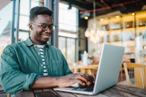 Young man sitting in coffee shop and using laptop. Shot of handsome young man sitting in city cafe and using laptop computer. black man laptop stock pictures, royalty-free photos & images