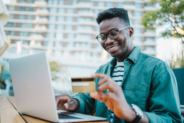 Man using laptop and credit card for online shopping in city cafe. Shot of young man doing shopping online with laptop and payment by credit card. Cheerful man using laptop computer and credit card in a sidewalk cafe. Student app stock pictures, royalty-free photos & images