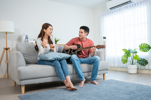 Asian young loving couple play guitar together in living room at home. Attractive romantic new marriage, active romantic man sitting on sofa and teach beautiful girlfriend play an instrument at home.
