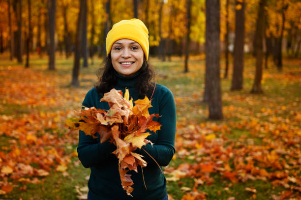 秋の黄色いオレンジ色の色とりどりの落ち葉の乾いた花束を手に、カメラを見つめる歯の生えた笑顔、黄金の秋の背景にポーズをとった陽気な女性のライフスタイルの肖像画 - autumn women smiling leaf ストックフォトと画像