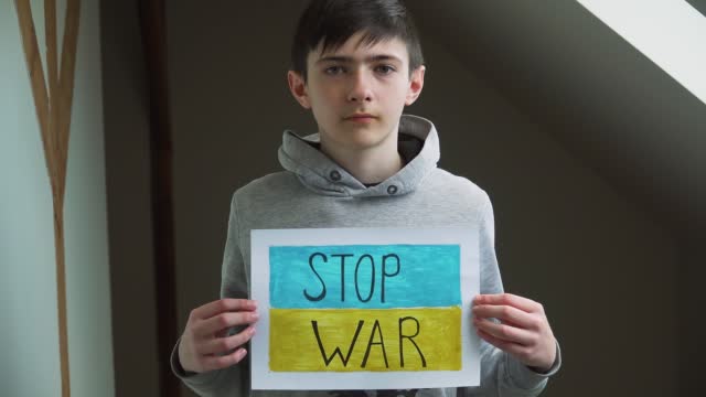 Boy holding self made picture with text Stop war on background of Ukrainian flag in refugee camps