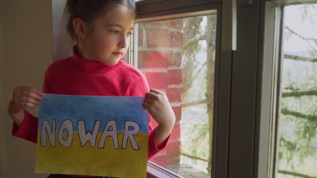 Refugee girl looking outside window and holding banner with inscription No war at blue yellow flag of Ukraine