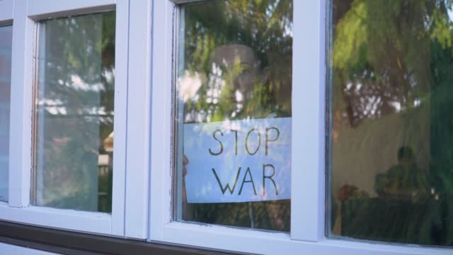 Scared woman showing through window simple placard with inscription Stop war