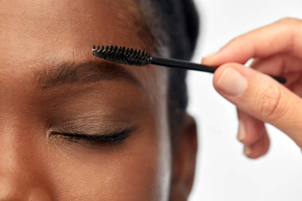 face of african woman and hand with mascara brush beauty, make up and cosmetics concept - close up of face of young african american woman and hand with mascara brush applying eyebrow shadows over white background lash and brow comb stock pictures, royalty-free photos & images