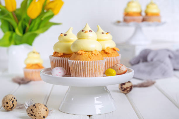 istock Cute Easter chick cupcakes. Vanilla cupcakes with buttercream on a white stand with fresh flowers on a white wooden background. Copy space. 1387068408