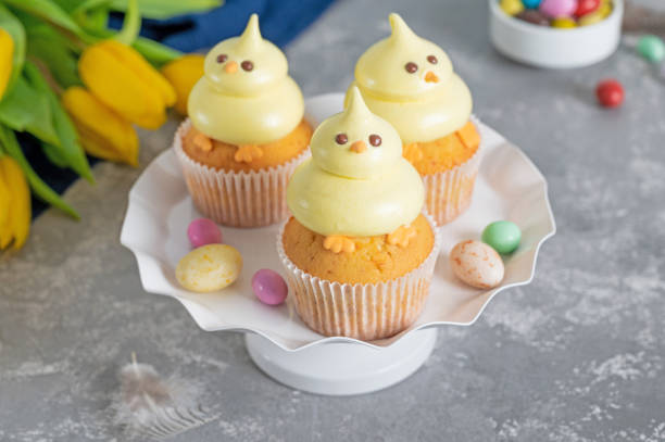 istock Cute Easter chick cupcakes. Vanilla cupcakes with buttercream on a white stand with fresh flowers on a white wooden background. Copy space. 1387068409