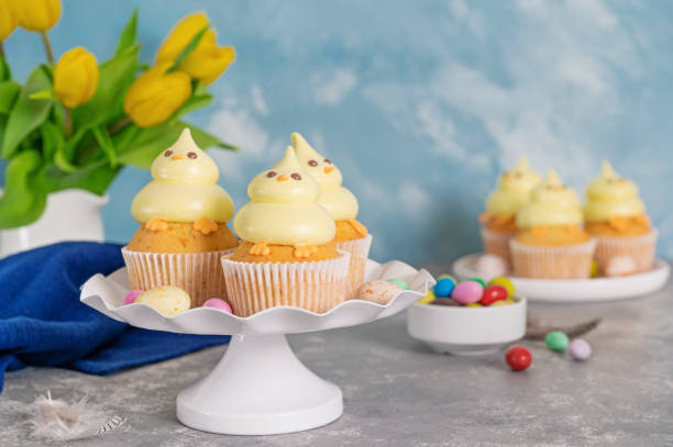 istock Cute Easter chick cupcakes. Vanilla cupcakes with buttercream on a white stand with fresh flowers on a white wooden background. Copy space. 1387068411