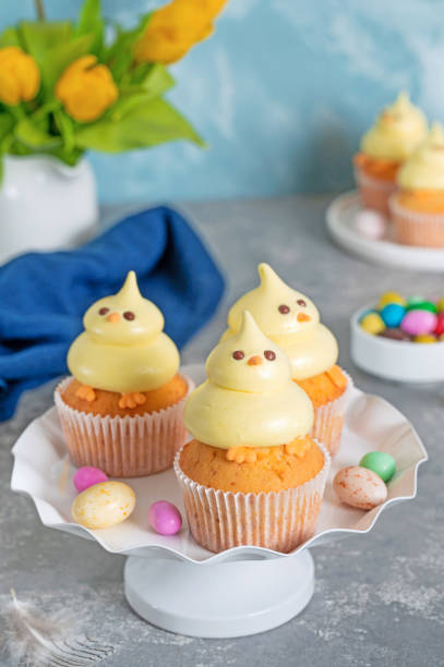 istock Cute Easter chick cupcakes. Vanilla cupcakes with buttercream on a white stand with fresh flowers on a white wooden background. Copy space. 1387068418