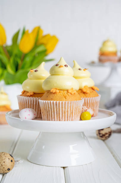 istock Cute Easter chick cupcakes. Vanilla cupcakes with buttercream on a white stand with fresh flowers on a white wooden background. Copy space. 1387068421