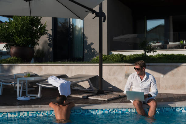 A modern elderly man enjoys the pool while working on his laptop next to a modern luxury house. Selective focus. High-quality photo