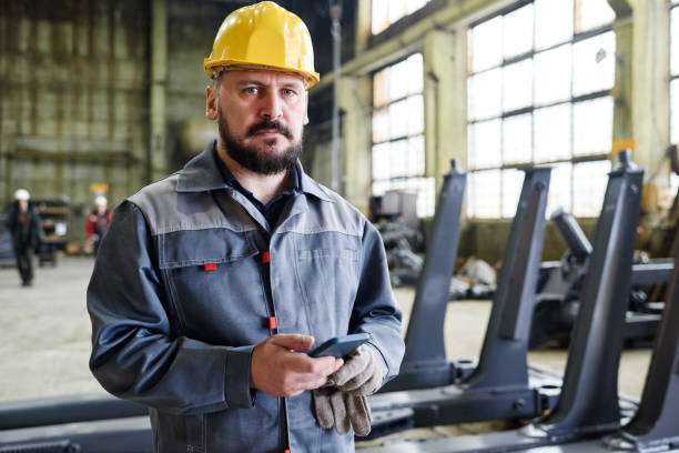 istock Serious mature workman or technician with smartphone in hardhat and boilersuit 1387074602