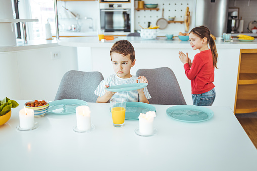 Little brother and sister helping to set the table at lunch time. Parents preparing meal in the background. Everybody is casually dressed, boy is 4 and girl is 5 years old. Horizontal waist up indoors shot with copy space.