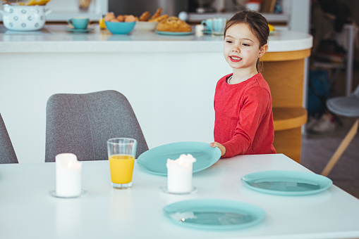 A young girl helps to set the family dining room table. Preschooler kid helping mother to set table in kitchen, carrying the plates, kitchen interior.