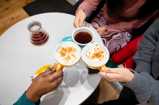 A high angle, close-up shot of three unrecognisable young women wearing warm casual clothing in a coffee shop on a winter's day. They are sitting and making a celebratory toast with their coffees.