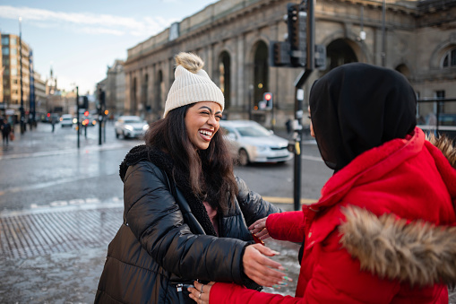 A wide shot of two young women wearing warm casual clothing, accessories and one of the women is wearing a hijab on a sunny winter's day in Newcastle Upon Tyne. They are meeting and embracing each other on the sidewalk of a city street.