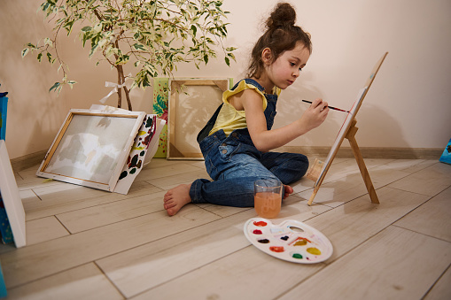 Adorable little Caucasian girl, concentrated artist, sitting on the floor next to a wooden easel and drawing picture, using watercolor paints. Kids art, entertainment, hobby and education concept.