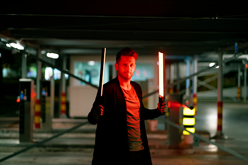 Portrait of a young Caucasian man at 
a parking lot of a garage holding two neon light sticks in his hands.