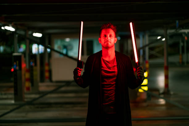 A front view of a serious young man holding two neon light sticks in both of his hands.