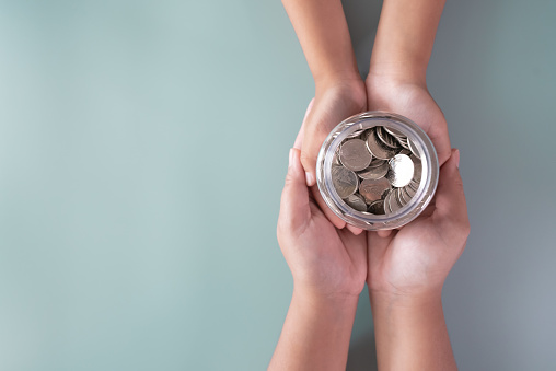 Adult and kid hands holding a coins jar together in savings and donation concept. Mother and child saving money together for donation, education, and future concept.