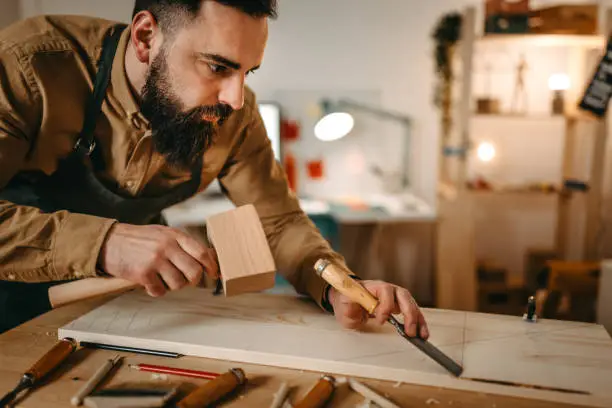 Focused young bearded artist carving wood with chisel and hammer on workbench in art studio