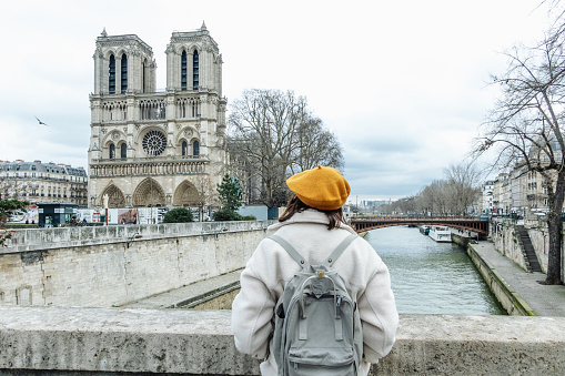 Photo of a young woman looking at Notre-Dame Cathedral and exploring the city on a cold, gloomy day; traveling to Paris alone, during the off-season.