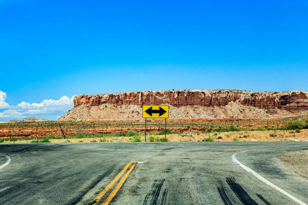 A Utah, USA perpendicular T-shaped crossroad road intersection with mid-distance flat prairie desert and a landslide rock formation butte in the far distance. A bright yellow directional arrow road sign informs motorists that either a full left or a full right turn are the only choices to continue traveling.