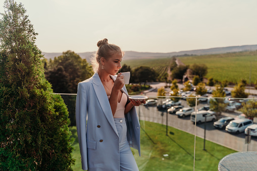 A middle-aged woman stands in a street cafe overlooking the mountains at sunset. She is wearing a blue jacket and drinking coffee while admiring nature. Travel and vacation concept