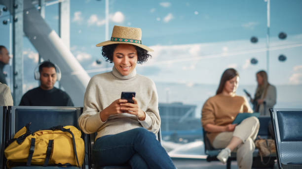 Airport Terminal: Happy Black Woman Waits for Flight, Uses Smartphone, Browse Internet, Post on Social Media, Online Shopping. Traveling African American Female in a Boarding Lounge of Airline Hub terminal del aeropuerto: la mujer negra feliz espera el vuelo, usa el teléfono inteligente, navega por internet, publica en las redes sociales, compras en línea. mujer afroamericana viajera en una sala de embarque del centro de operaciones de la aerolín