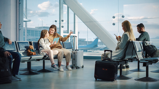 Airport Airplane Terminal: Cute Mother and Little Daughter Wait for their Vacation Flight, Play e-Learning e-Educational Video Games on Digital Tablet. Young Family in Boarding Lounge of Airline Hub
