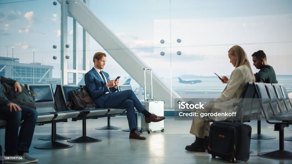 Busy Airport Terminal: Handsome Businessman Uses Smartphone While Waiting for His Flight. People Sitting in a Boarding Lounge of Big Airline Hub with Airplanes Departing and Arriving Airport Stock Photo