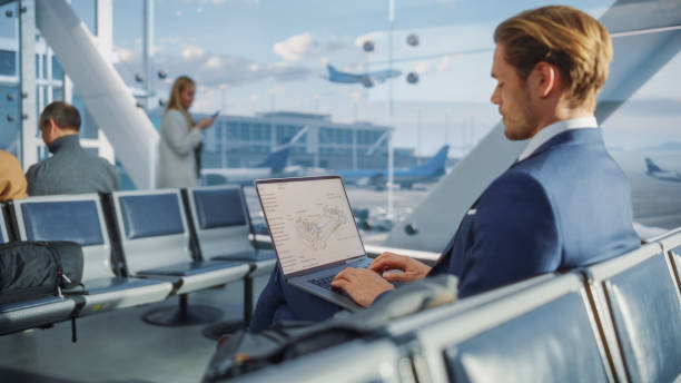 istock Airport Terminal: Businessman is Reviewing a Blueprint on Laptop While Waiting for His Plane Flight. Entrepreneur Does Online Remote Work in Boarding Lounge of Airline Hub. Over the Shoulder 1387097577