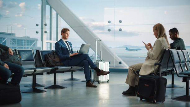 istock Busy Airport Terminal: Handsome Businessman Working on Laptop Computer While Waiting for His Flight. People Sitting in a Boarding Lounge of Big Airline Hub with Airplanes Departing and Arriving 1387097708