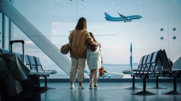 空港ターミナル:美しい母とかわいい娘は、到着し、飛行機を出発するための窓の外を見て、彼らの休暇のフライトを待っています。航空会社ハブの搭乗ラウンジの若い家族 - concepts airport ideas watching ストックフォトと画像