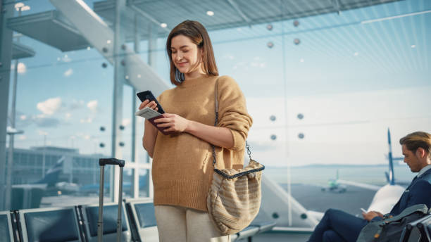 Airport Terminal: Happy Traveling Caucasian Woman Waiting at Flight Gates for Plane Boarding, Uses Mobile Smartphone, Checking Trip Destination on Internet. Smiling White Female on Vacation Trip terminal del aeropuerto: mujer caucásica feliz que viaja esperando en las puertas de vuelo para el embarque en avión, utiliza un teléfono inteligente móvil, verifica el destino del viaje en internet. mujer blanca sonriente en viaje de vacaciones