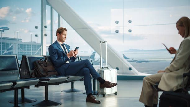 istock Airport Terminal: Businessman Uses Smartphone, Waiting for a Flight, Doing e-Business, Sending e-Commerce emails. Traveling Entrepreneur Remote Work Online Sitting in a Boarding Lounge of Airline Hub 1387098358